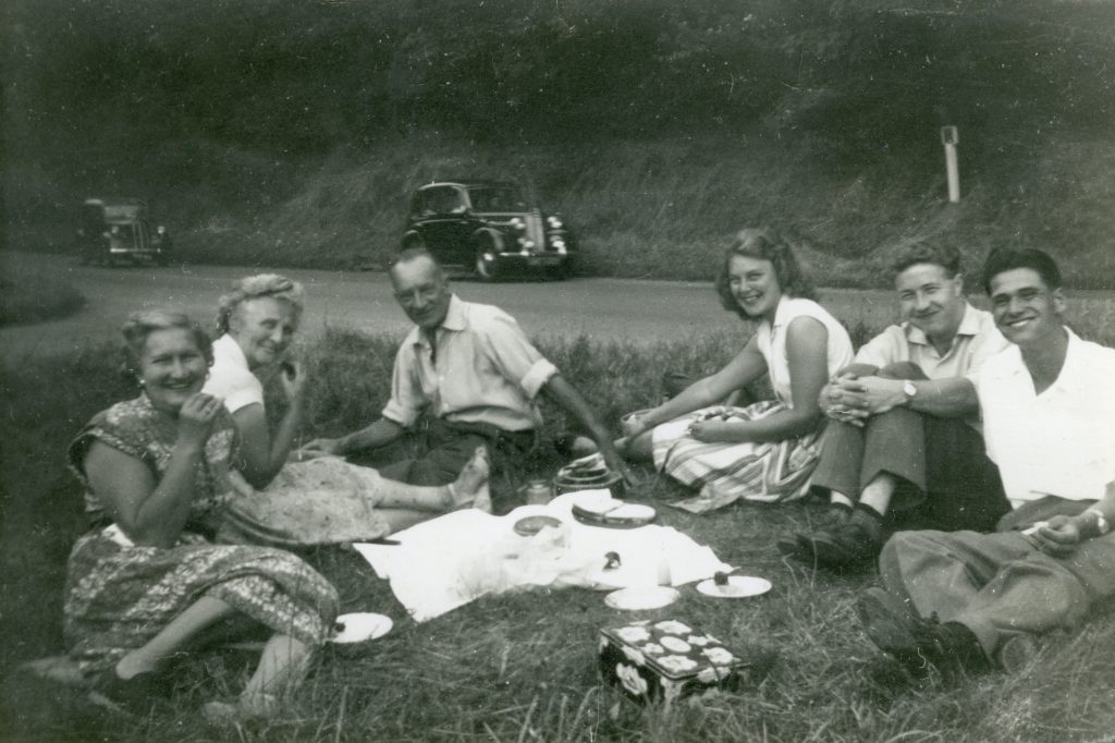 A vintage photo of a family having a picnic on the side of the road wth a car parked across the road.