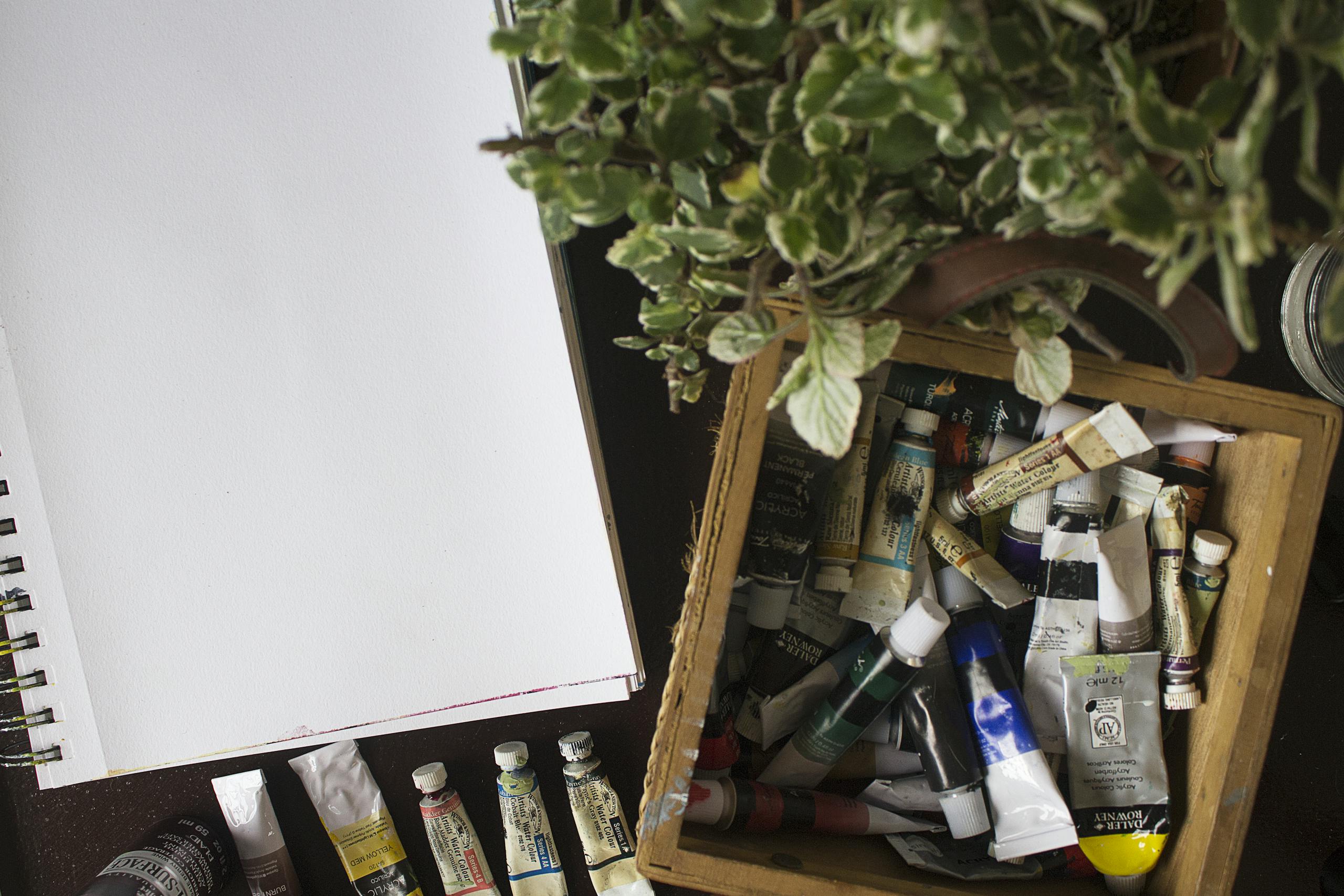 A variety of paint tubes next to a blank sketchbook, surrounded by greenery, inviting creativity.